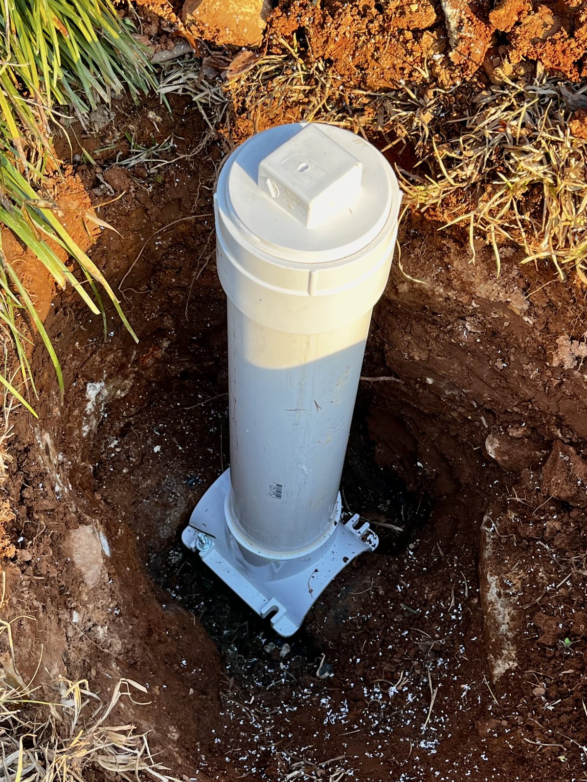 New PVC sewer cleanout riser with a white cap set in an excavated hole in red clay